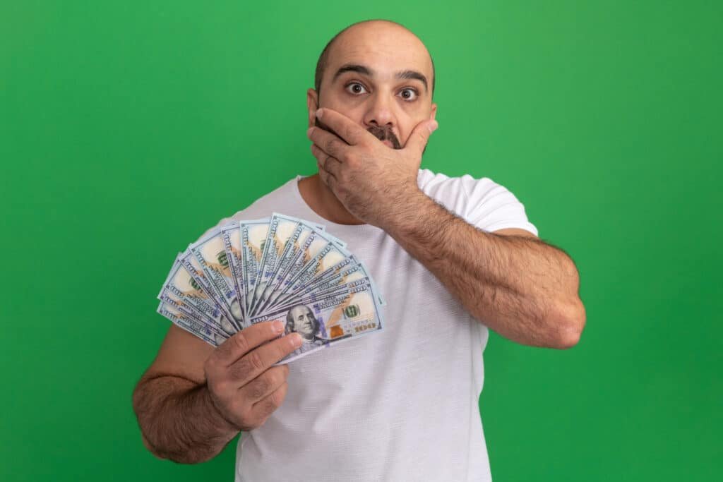 bearded man in white t shirt holding cash looking at camera being shocked covering mouth with hand standing over green background